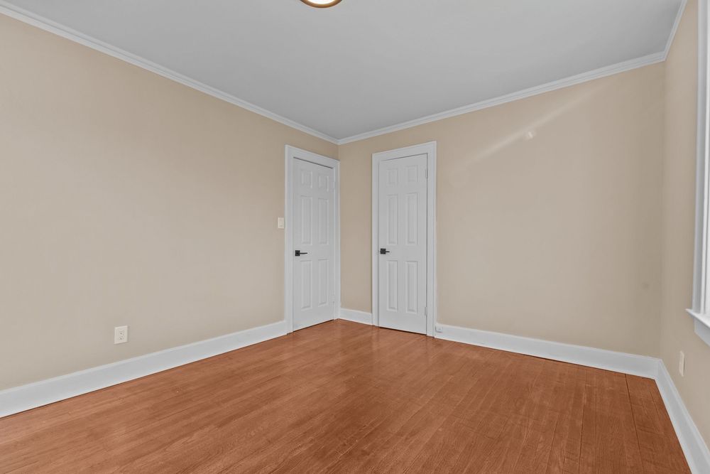 Empty bedroom with hardwood floors, crown molding, two white doors, and neutral beige walls in a clean bright space.