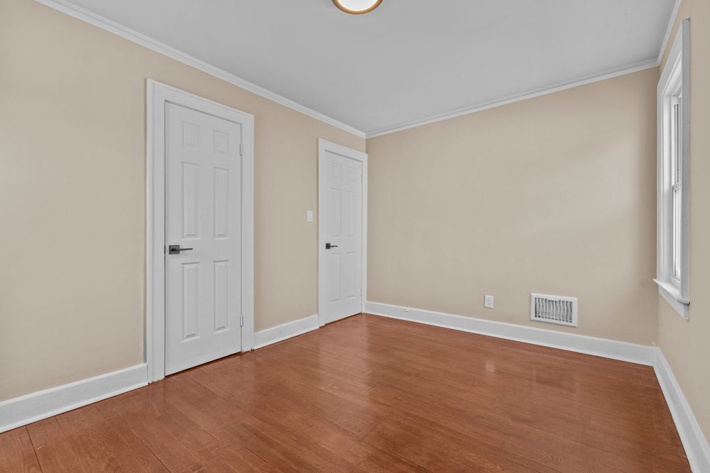 Empty bedroom with hardwood floors, beige walls, crown molding, two white doors, and a floor vent.