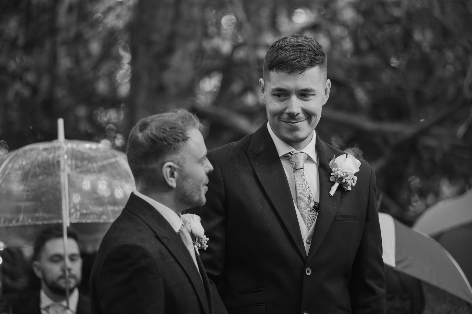 Black and white photo of two grooms smiling at outdoor wedding ceremony with umbrella visible in background.
