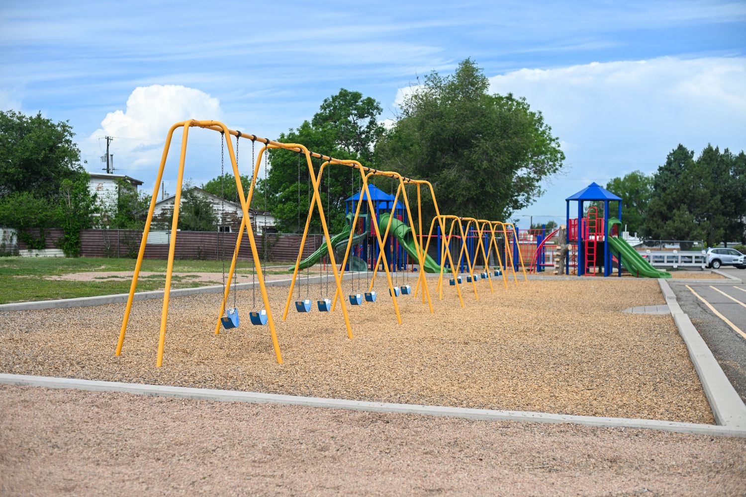 Colorful yellow swing set on wood chip playground with play structure and trees under blue sky.