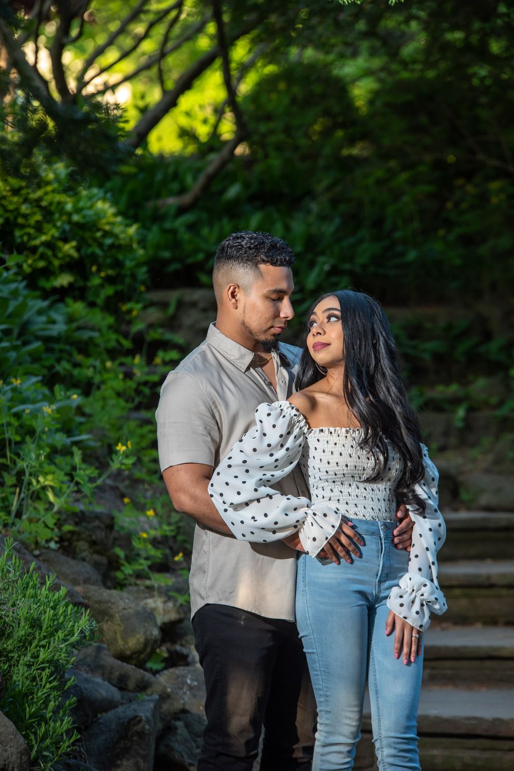 Romantic couple holding each other in a sunlit garden with stone steps behind them.