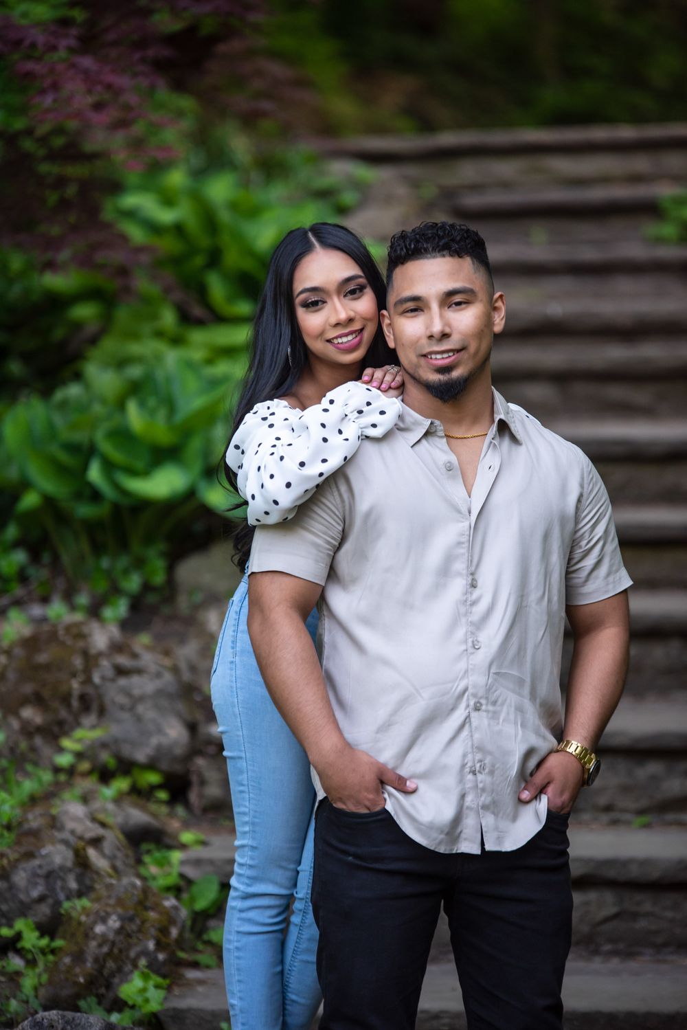 Couple posing together on stone garden steps surrounded by lush green foliage in a park setting.