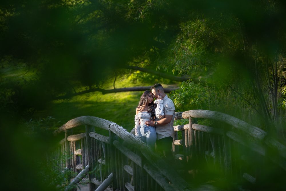 Couple kissing on a wooden bridge framed by lush green trees in soft golden sunlight.