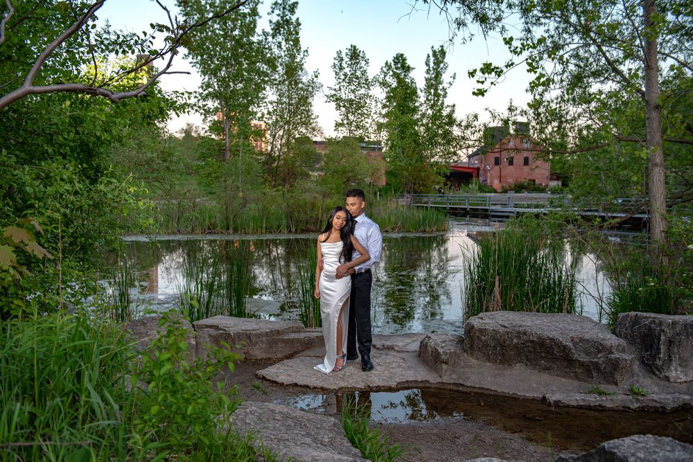 Couple standing on rocks by a serene pond surrounded by green trees at sunset.