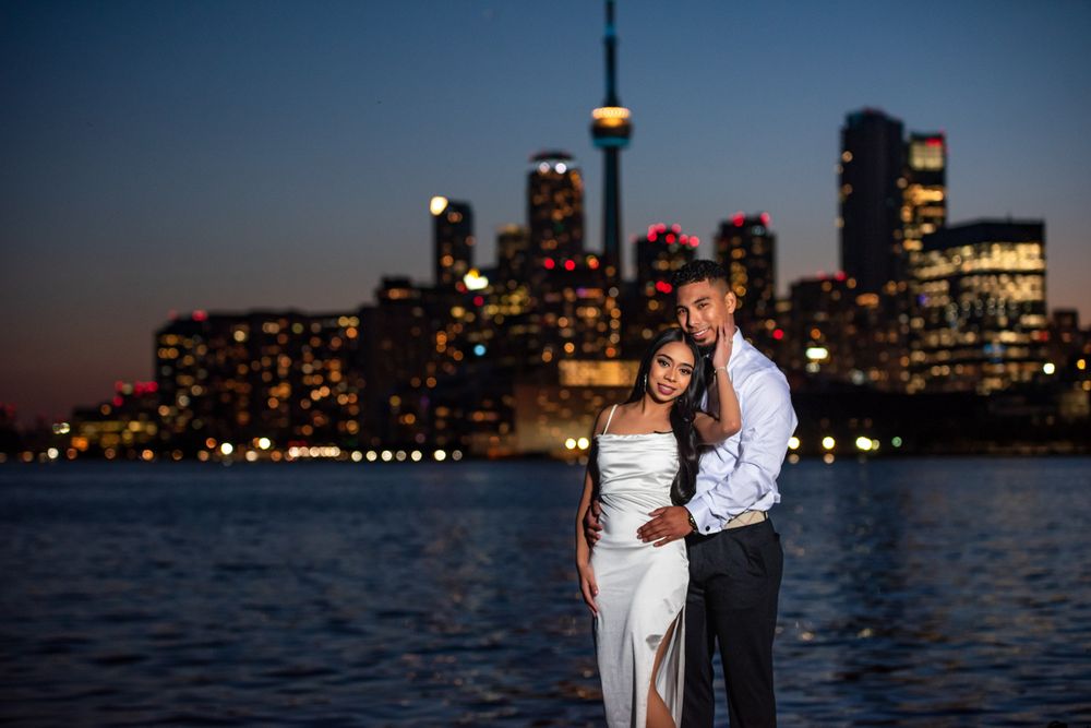 Romantic couple posing together at dusk with the glowing Toronto skyline and CN Tower visible behind them.