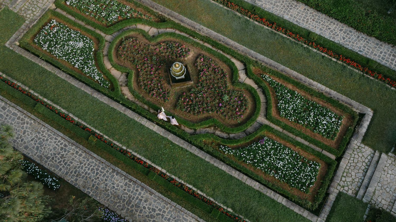 Aerial view of an ornate formal garden with geometric patterns, manicured hedges, colorful flowers, and a central decorative fountain.