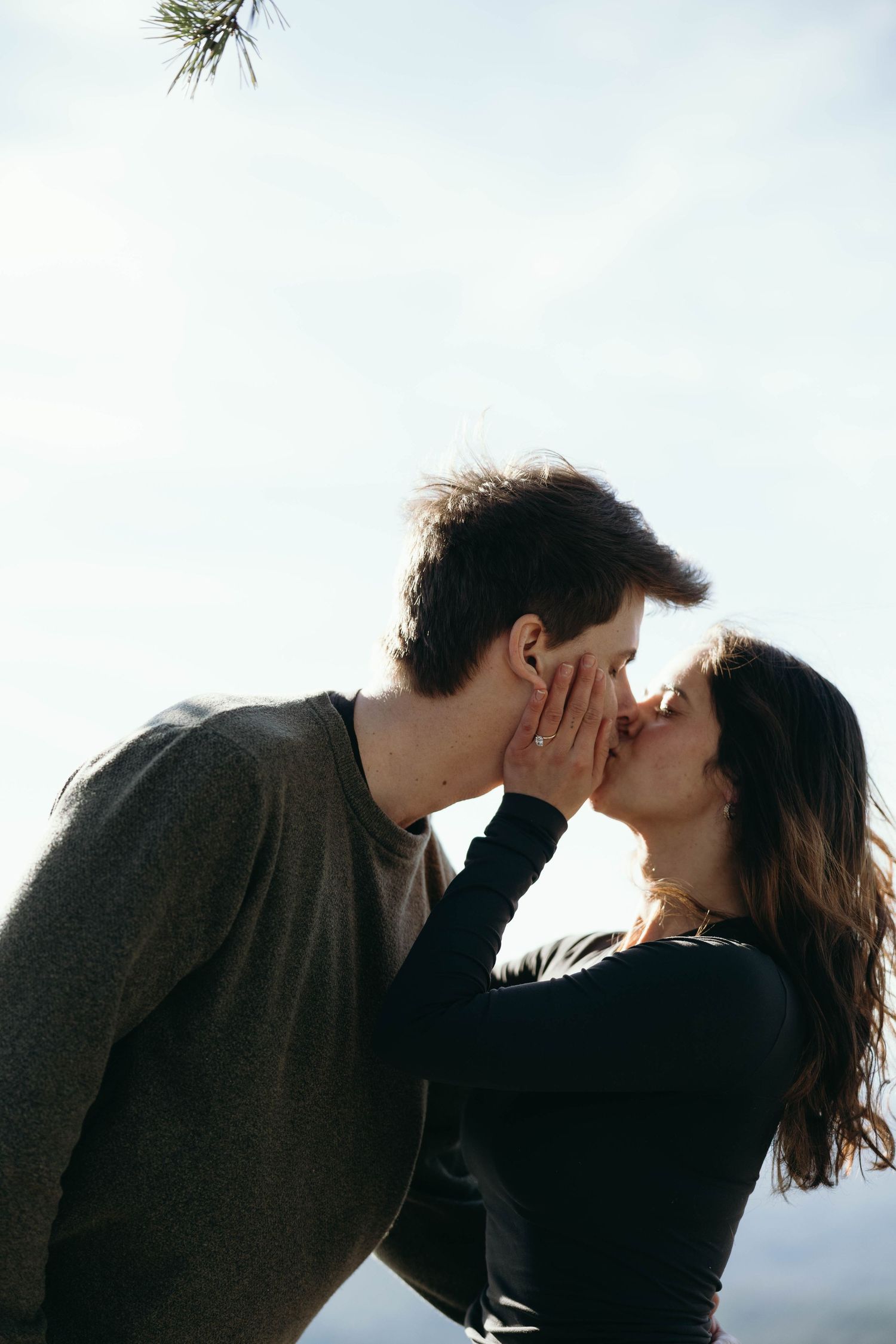 Romantic couple sharing a tender kiss outdoors against a bright sky, woman gently touching man's face.