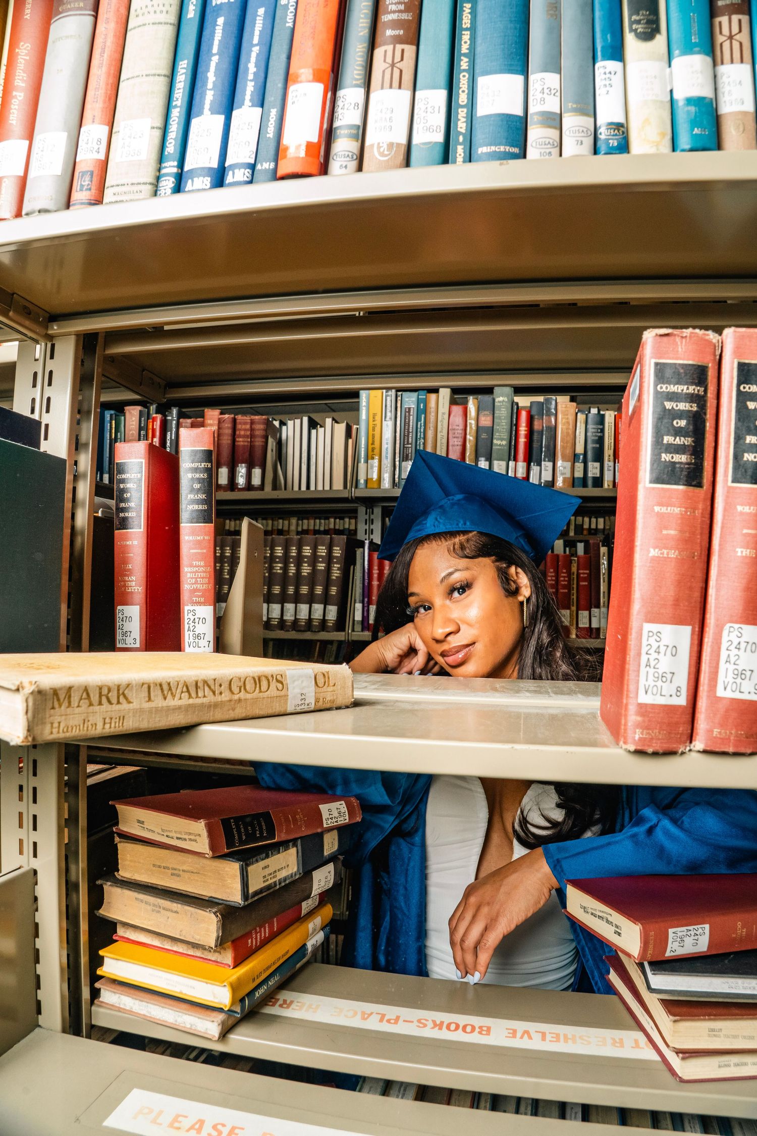 A young woman in blue graduation cap and gown poses between library bookshelves filled with books.
