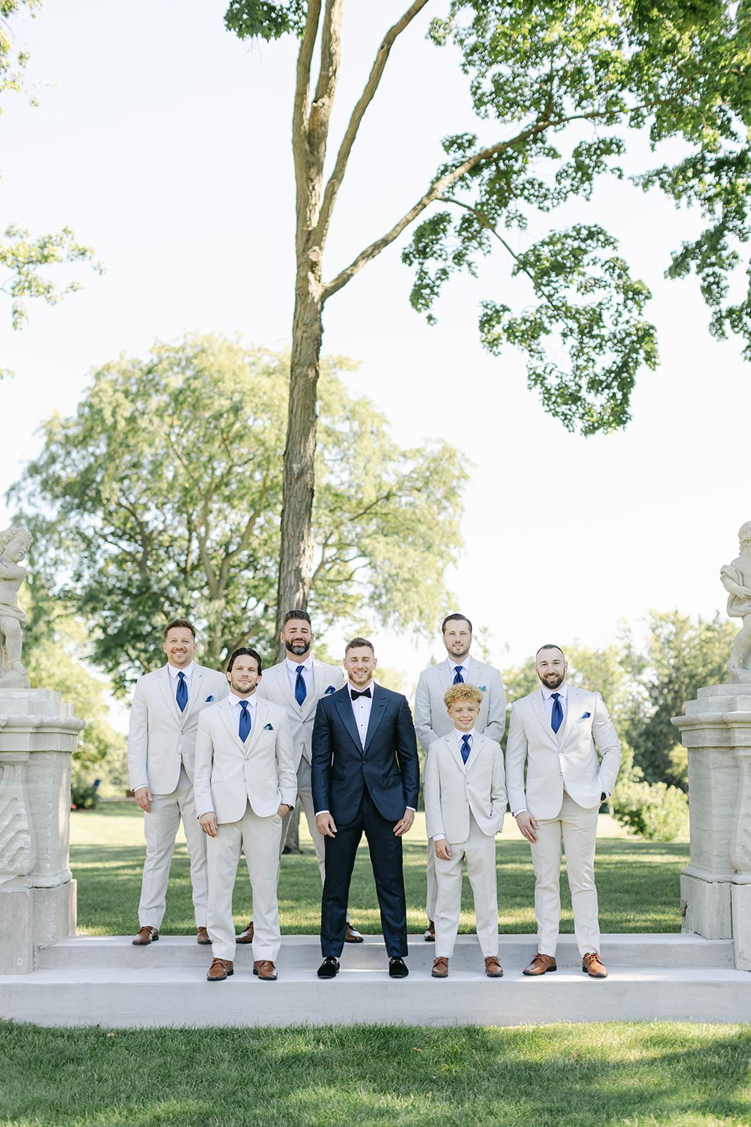 Armour House Wedding - Groom in navy tuxedo stands with groomsmen in tan suits and blue ties outdoors at a wedding ceremony.