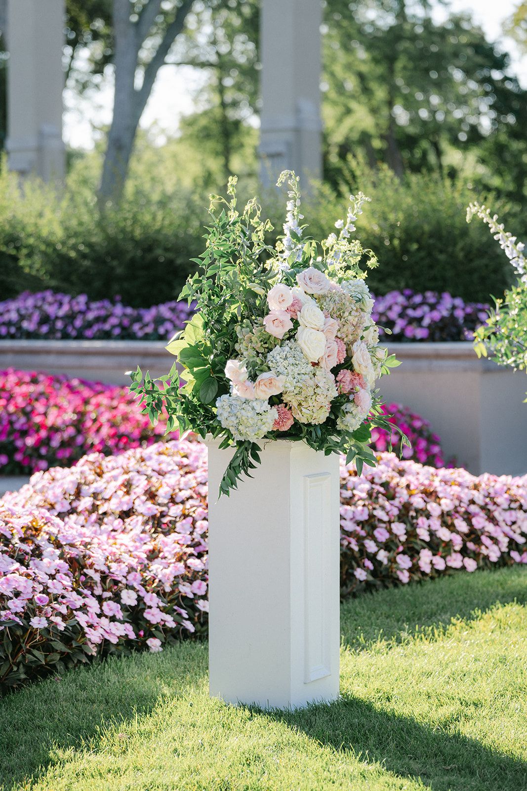 Armour House Wedding - White pedestal with lush floral arrangement of roses and hydrangeas, set against vibrant garden flower beds.