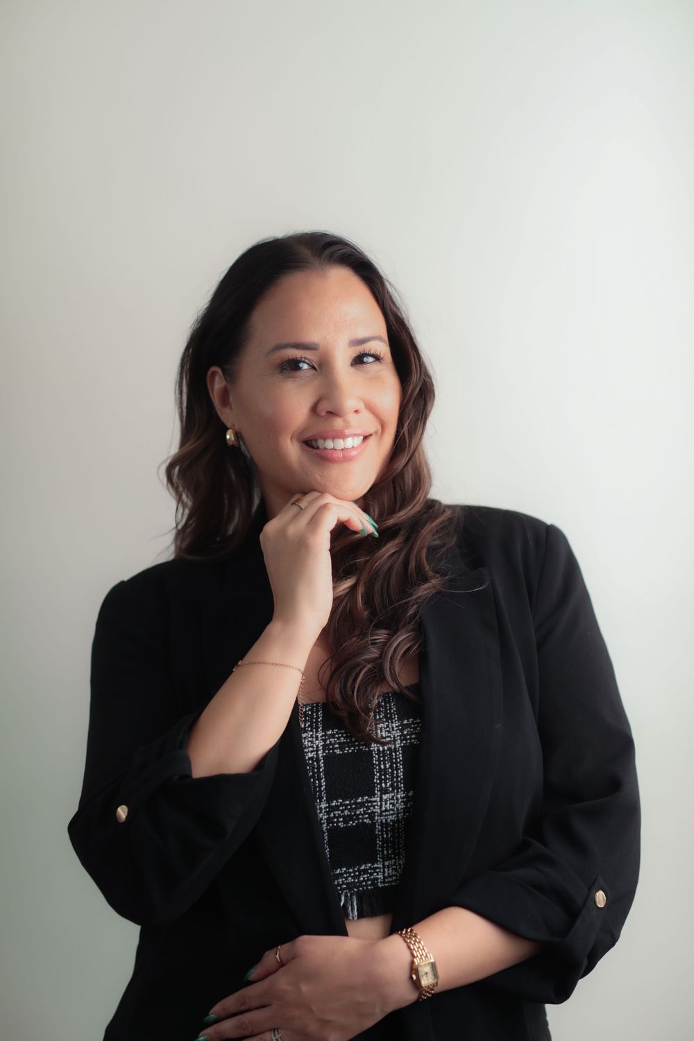 Professional woman in black blazer smiling confidently with hand on chin against light background.