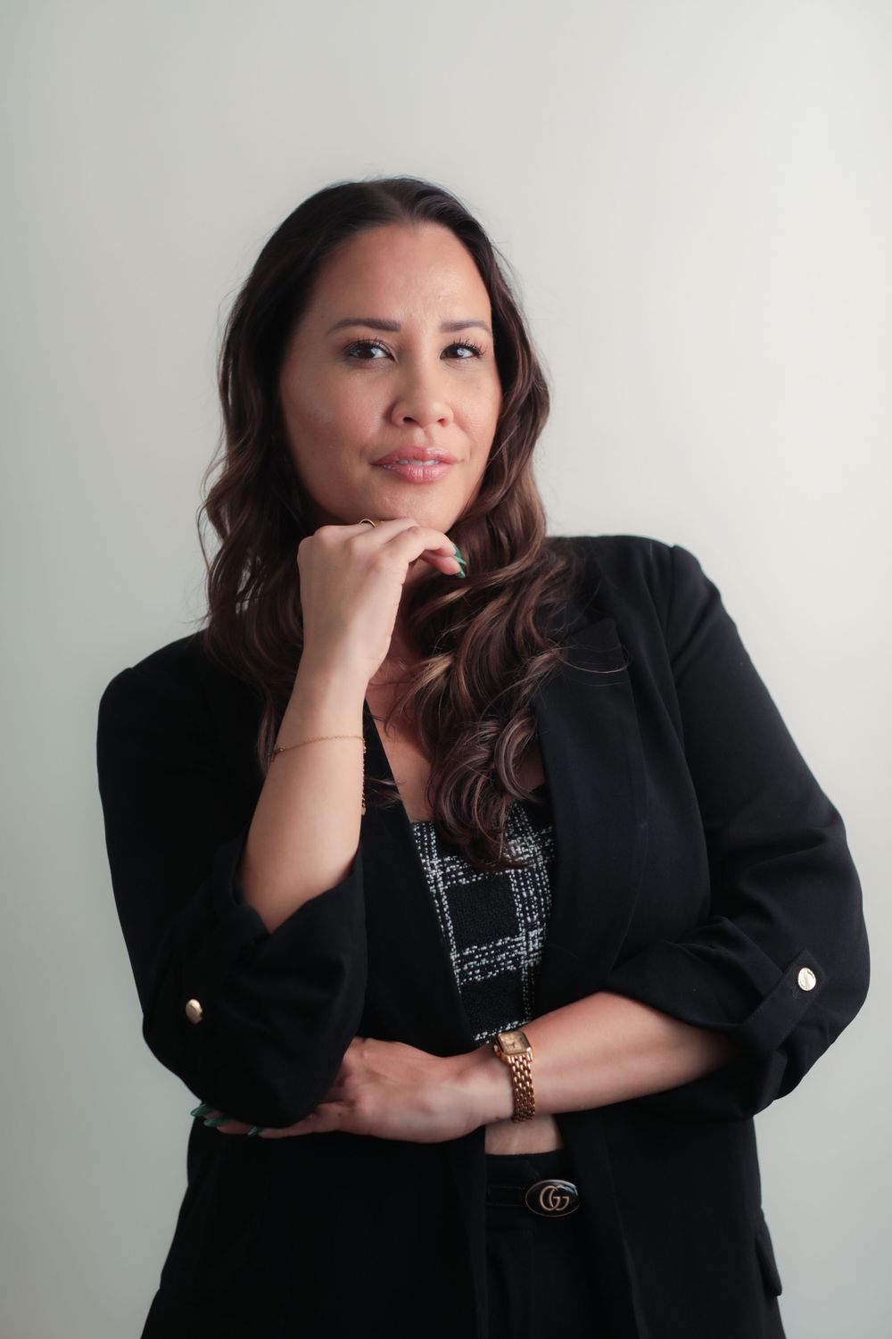 Professional woman in black blazer posing thoughtfully with hand on chin, wearing gold Gucci bracelet.