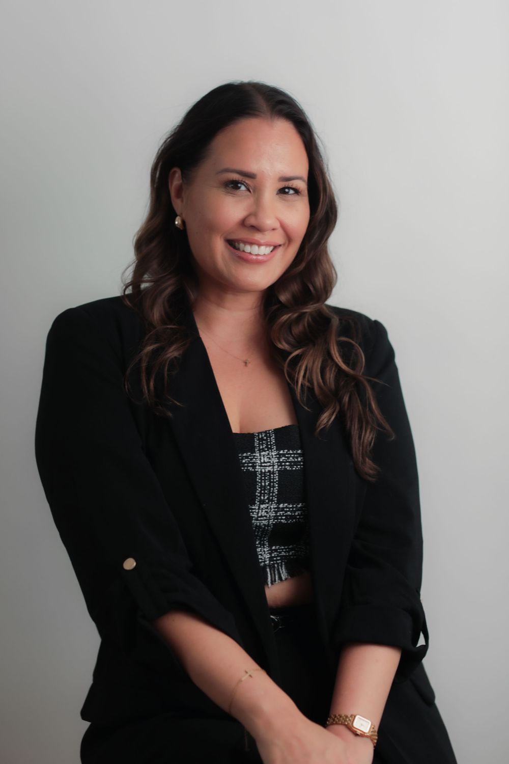 Professional woman in black blazer smiling confidently, wearing a gold watch, with long brown hair.