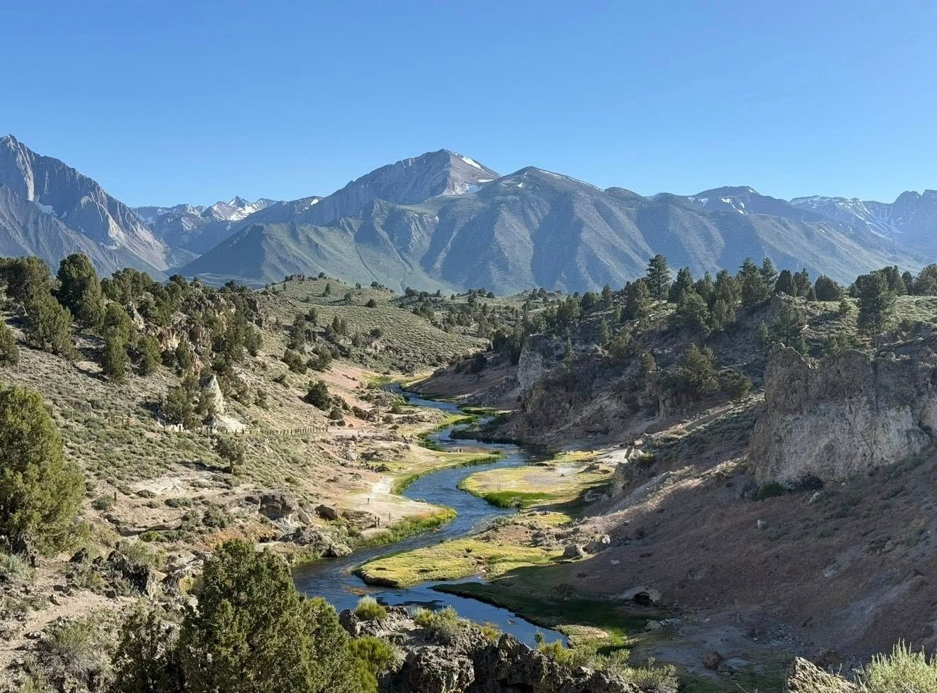 Scenic mountain valley with winding river, lush greenery, rocky terrain, and snow-capped peaks under clear blue sky.