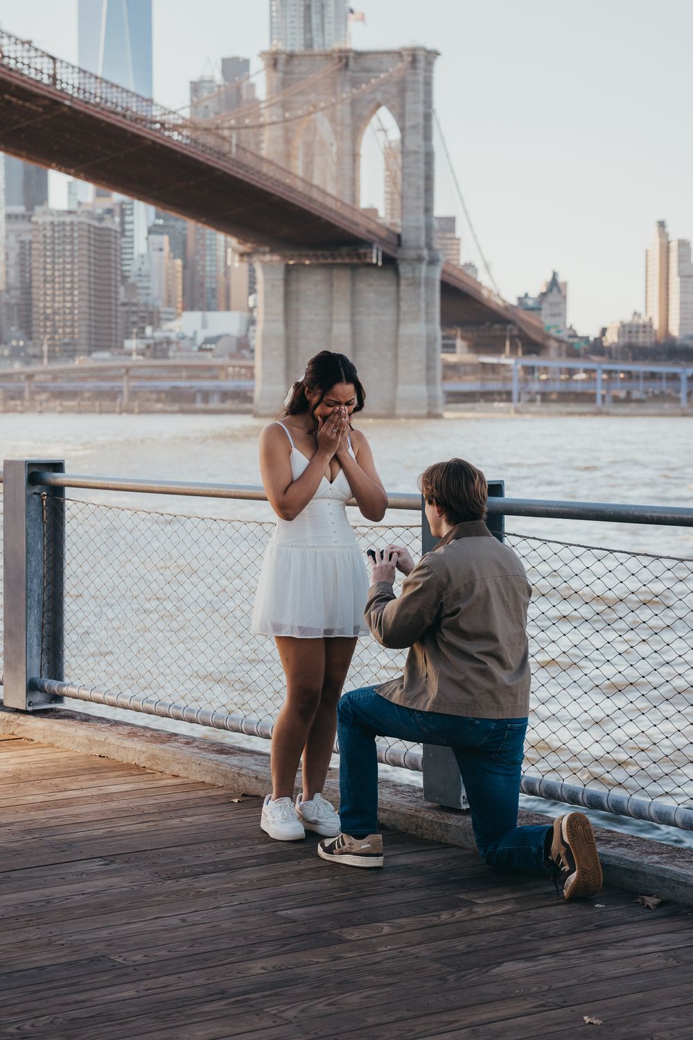 Man proposing on one knee to his surprised girlfriend on a Brooklyn Bridge waterfront dock.