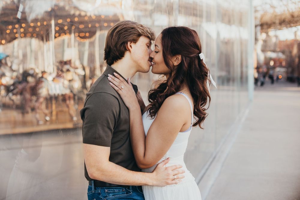 Young couple sharing a romantic kiss in front of a glowing carousel at sunset.