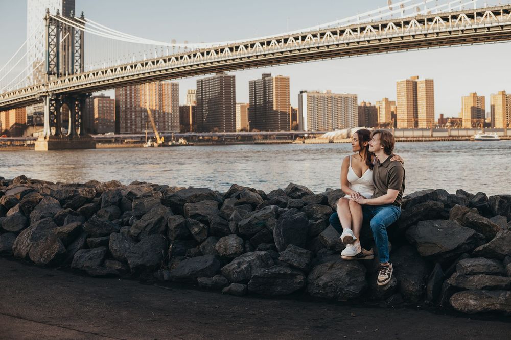 Couple sitting together on rocks with the Manhattan Bridge and NYC skyline at golden hour.