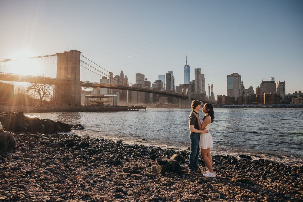 Couple kissing on a rocky shore with the Brooklyn Bridge and NYC skyline at sunset.