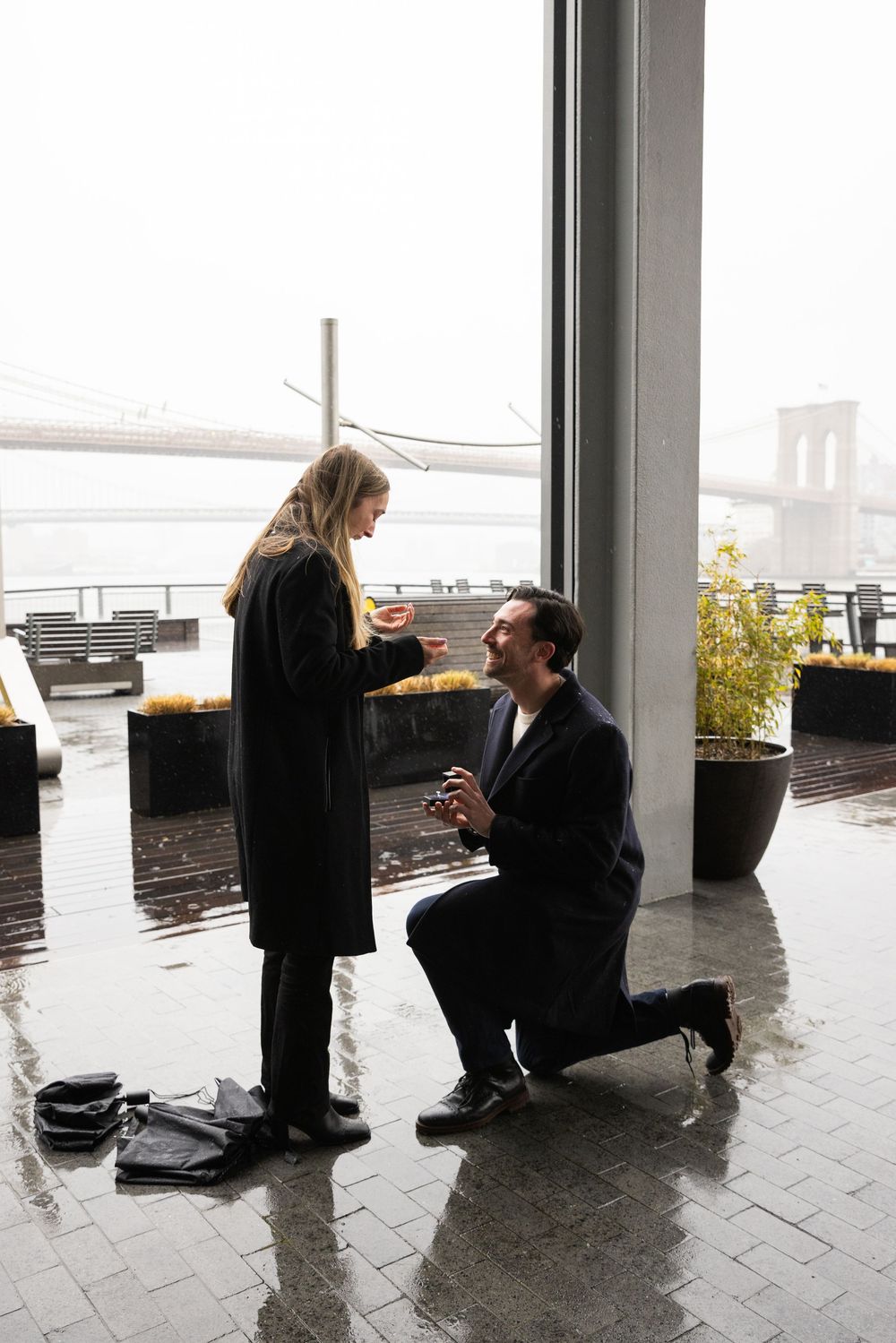 Man proposing on one knee to woman near a window with Brooklyn Bridge in the background.