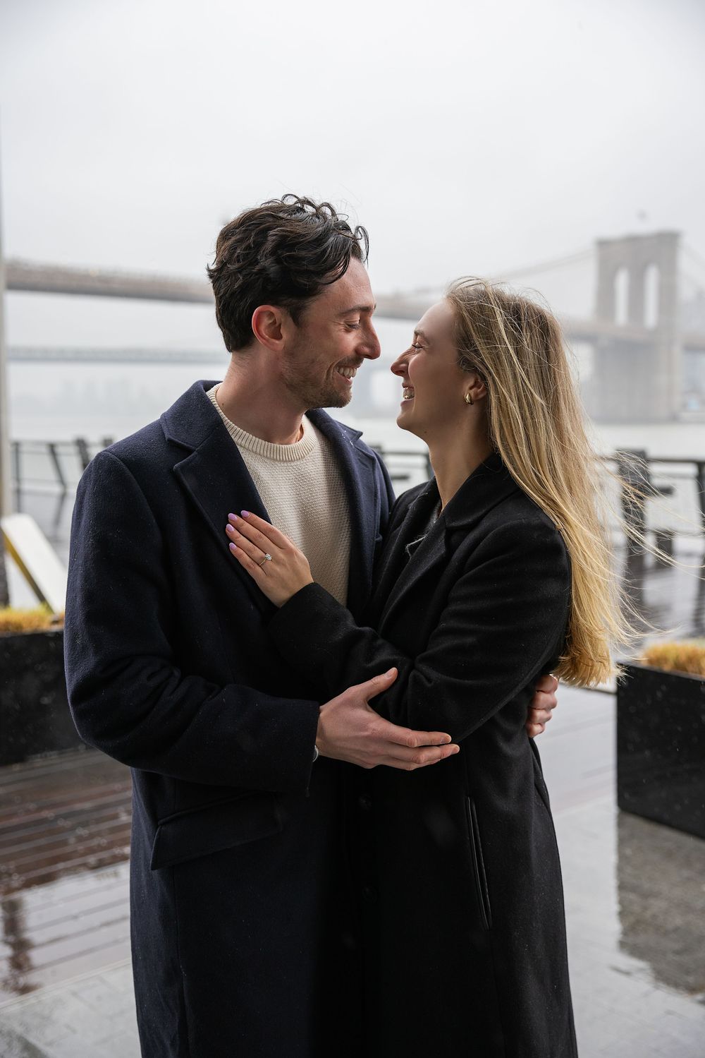 Smiling couple embracing outdoors near the Brooklyn Bridge on a rainy overcast day.