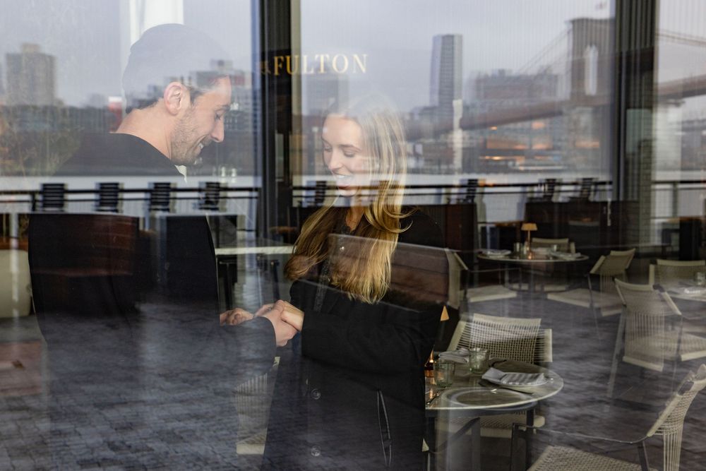 Couple sharing an intimate moment at Fulton restaurant with Brooklyn Bridge views through glass.