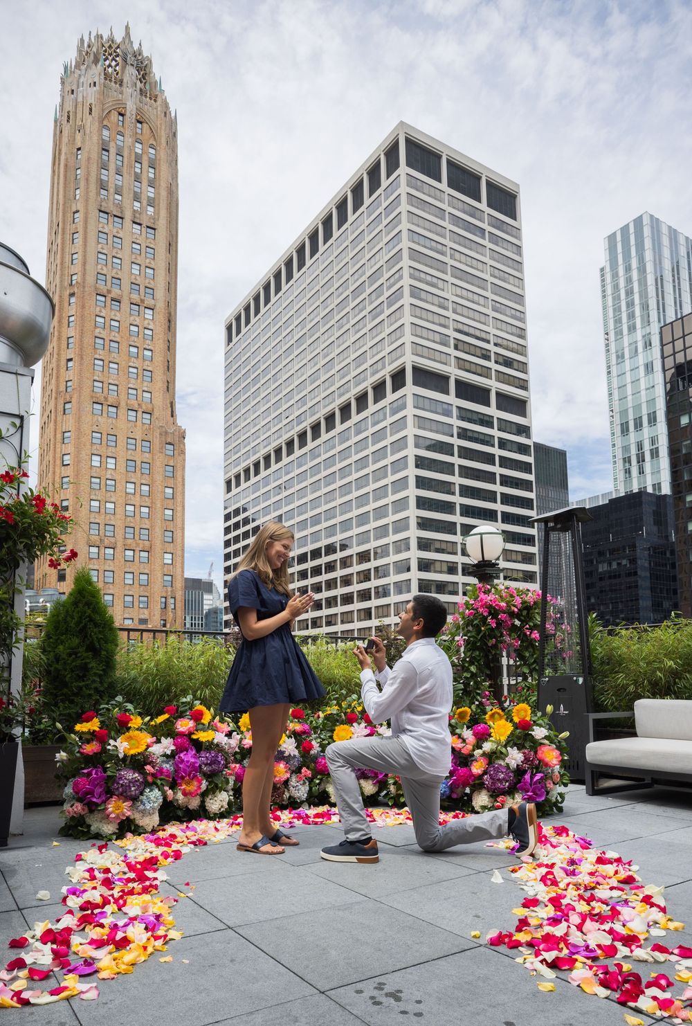 Man proposing on one knee to woman on a rooftop surrounded by rose petals and flowers.