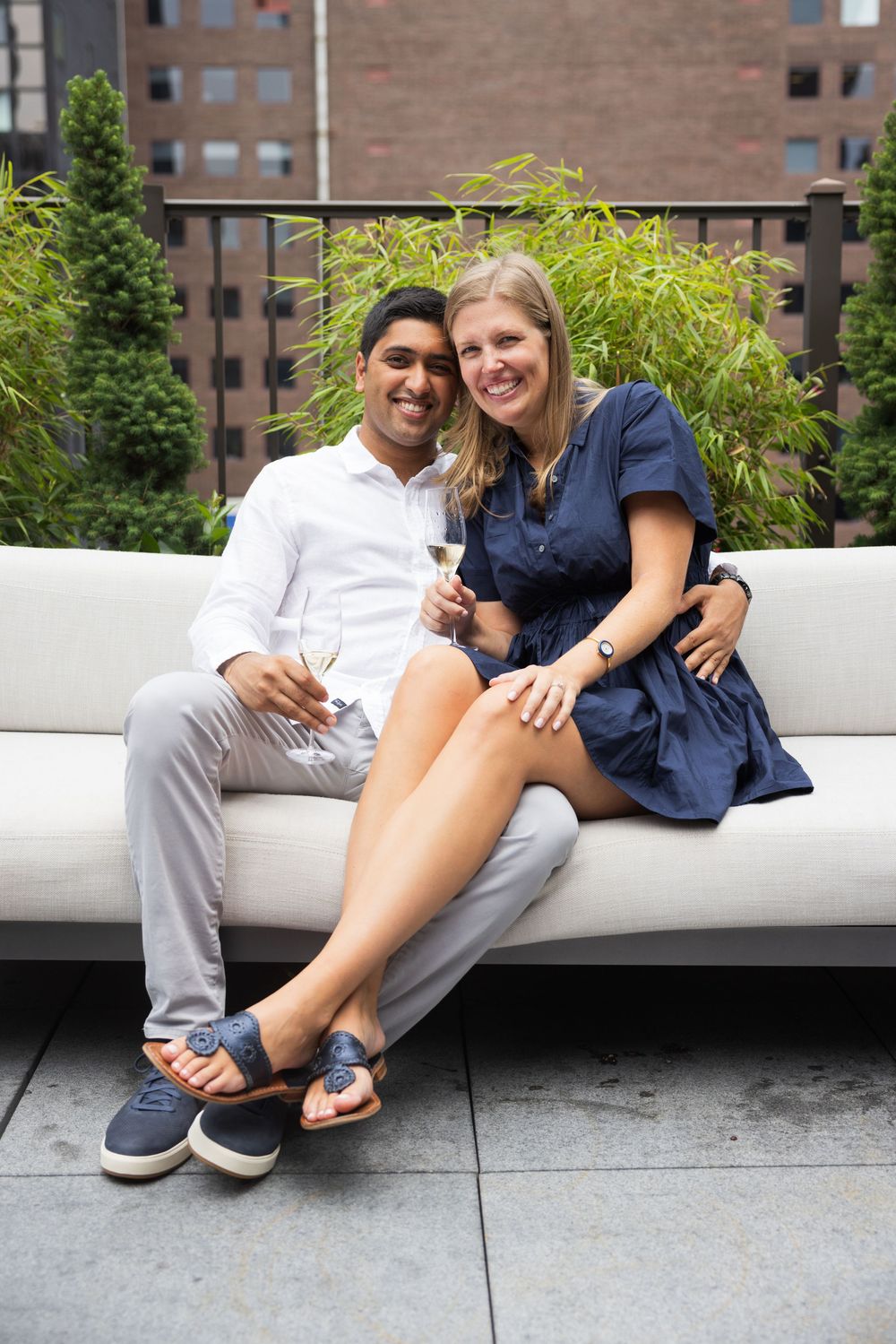 Happy couple sitting on a white outdoor sofa toasting with champagne glasses on a rooftop.