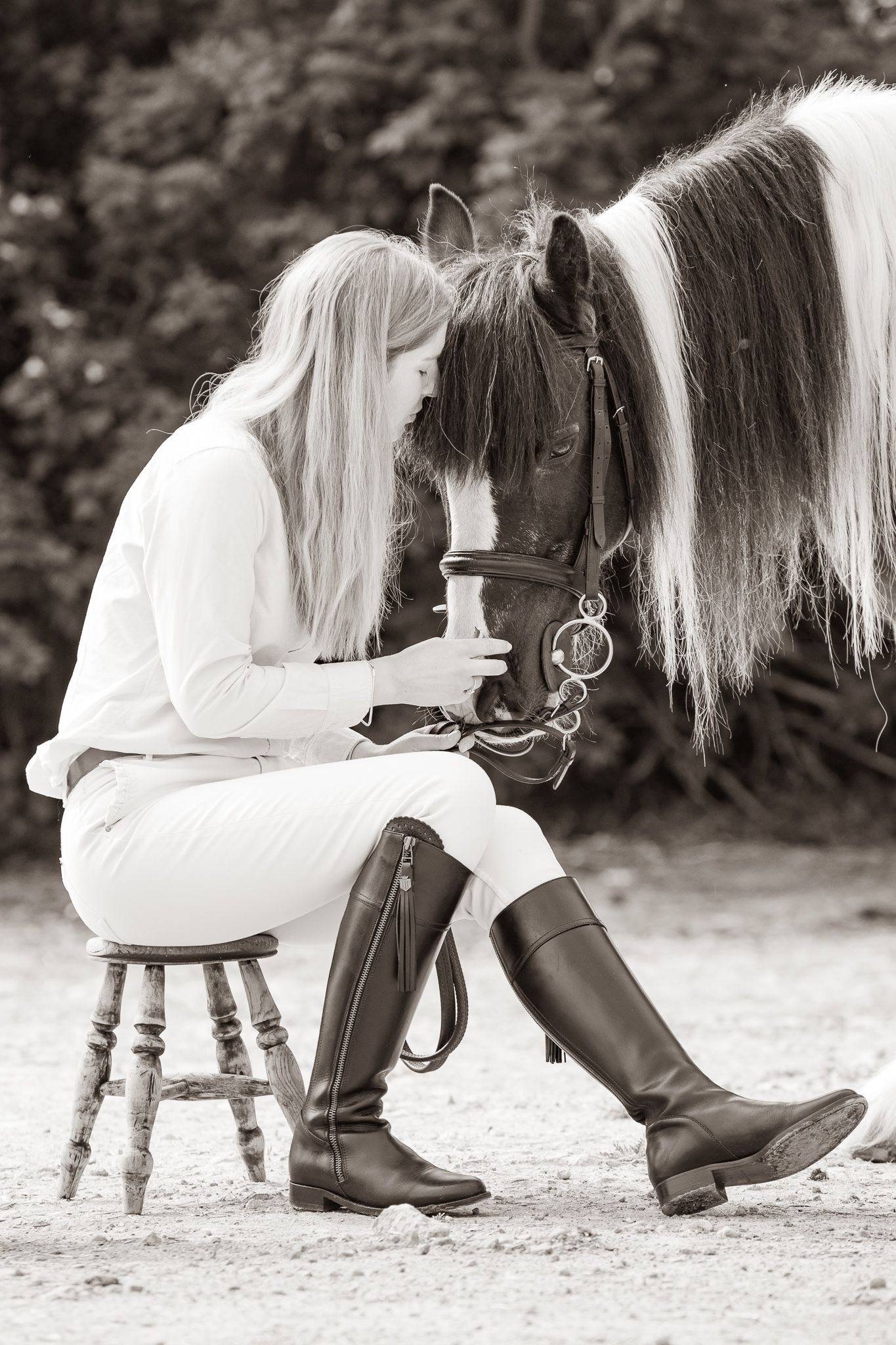 A beautiful bay horse wearing a black bridle stands next to someone in a white lace dress in an outdoor setting.