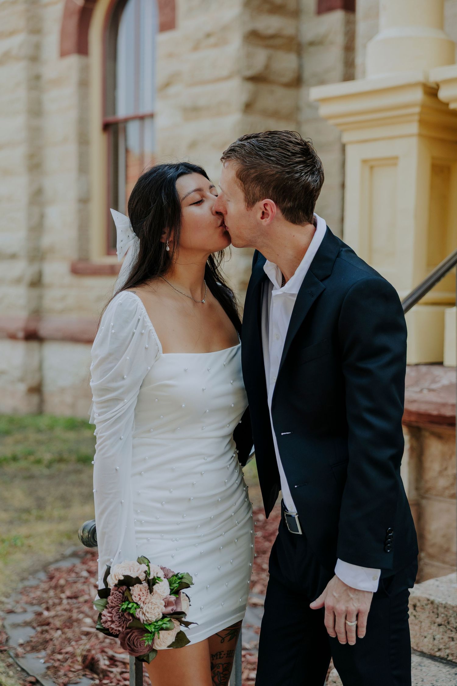 Couple kissing in front of Texas State Capitol building, bride raising bouquet, groom in navy suit, outdoor wedding photo.