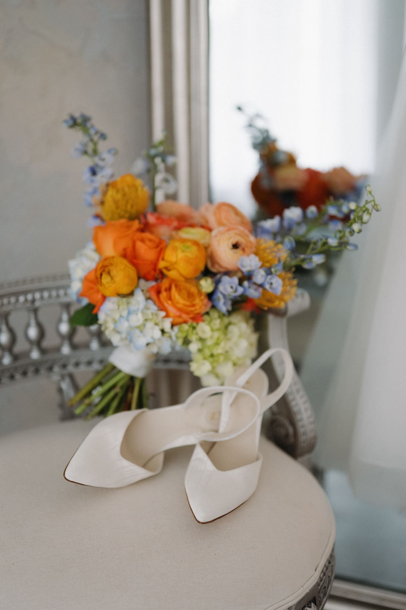 White bridal heels rest before a vibrant wedding bouquet of orange ranunculus and blue hydrangeas on an elegant table.