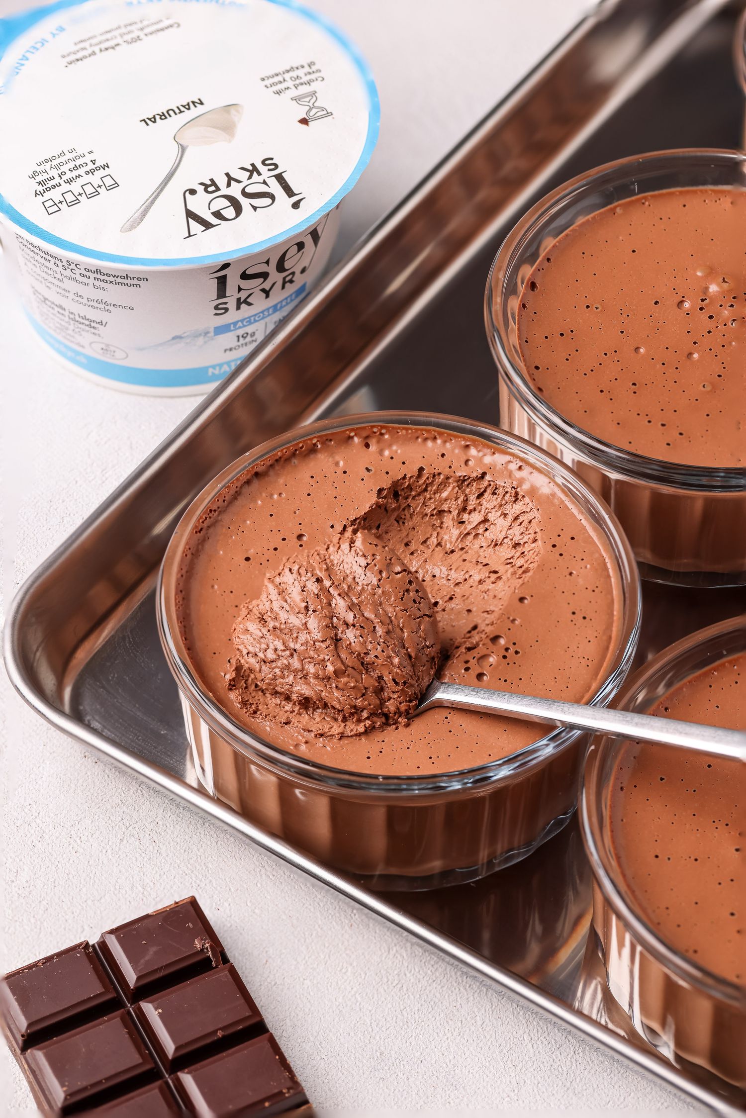 Chocolate mousse desserts in glass jars with a bar of dark chocolate on a white surface.