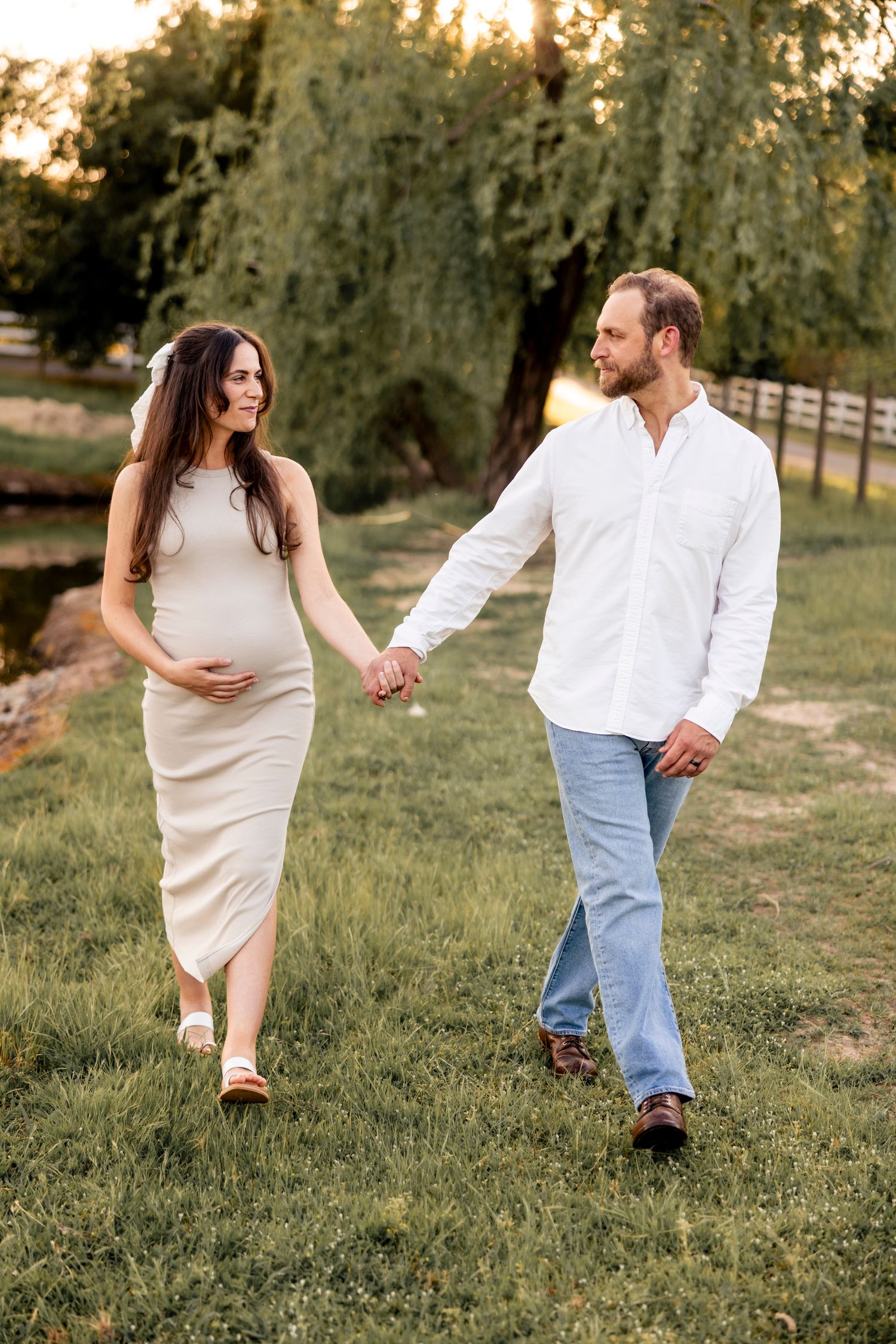 Pregnant woman in beige dress holds hands with man in white shirt walking on green grass near willow tree.