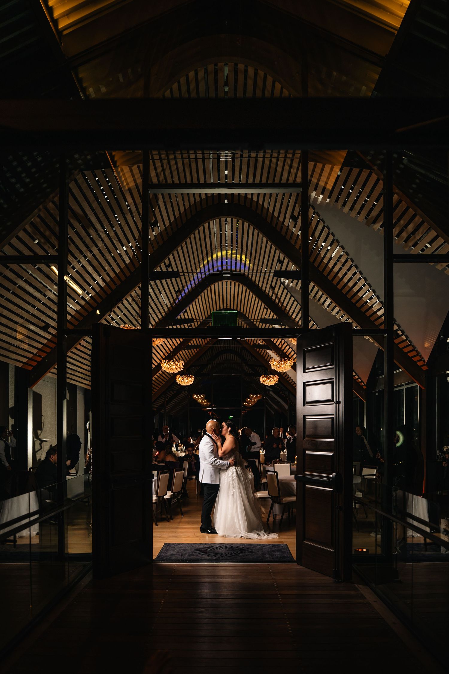 Couple sharing first dance kiss in elegant venue with arched wooden ceiling and warm ambient lighting.