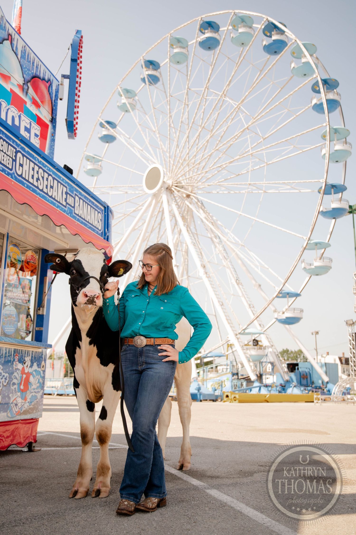 A young woman in a teal shirt and jeans stands with a Holstein cow at a county fair, with a Ferris wheel behind her.