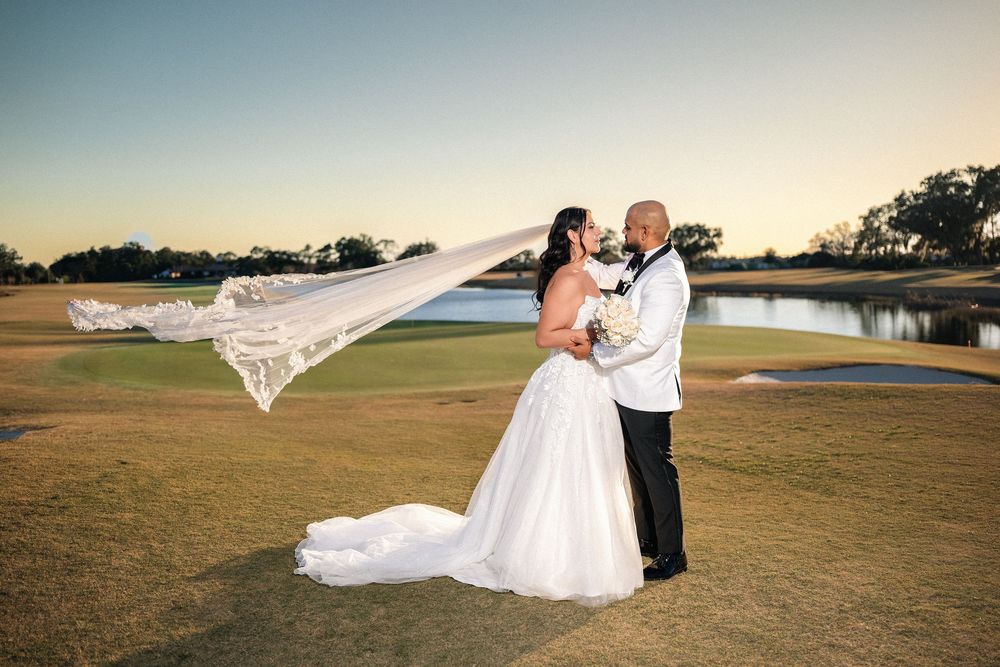 Bride and groom pose on golf course at sunset, her veil flowing in the wind, wearing white gown and tuxedo.