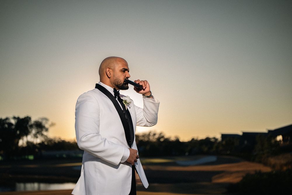Groom in white tuxedo sipping a drink at sunset outdoors during a wedding celebration.