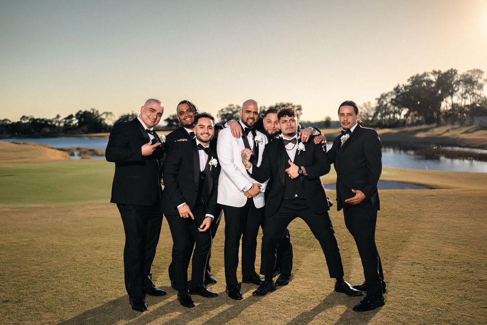 Groom and groomsmen in black tuxedos posing on a golf course at sunset during a wedding celebration.