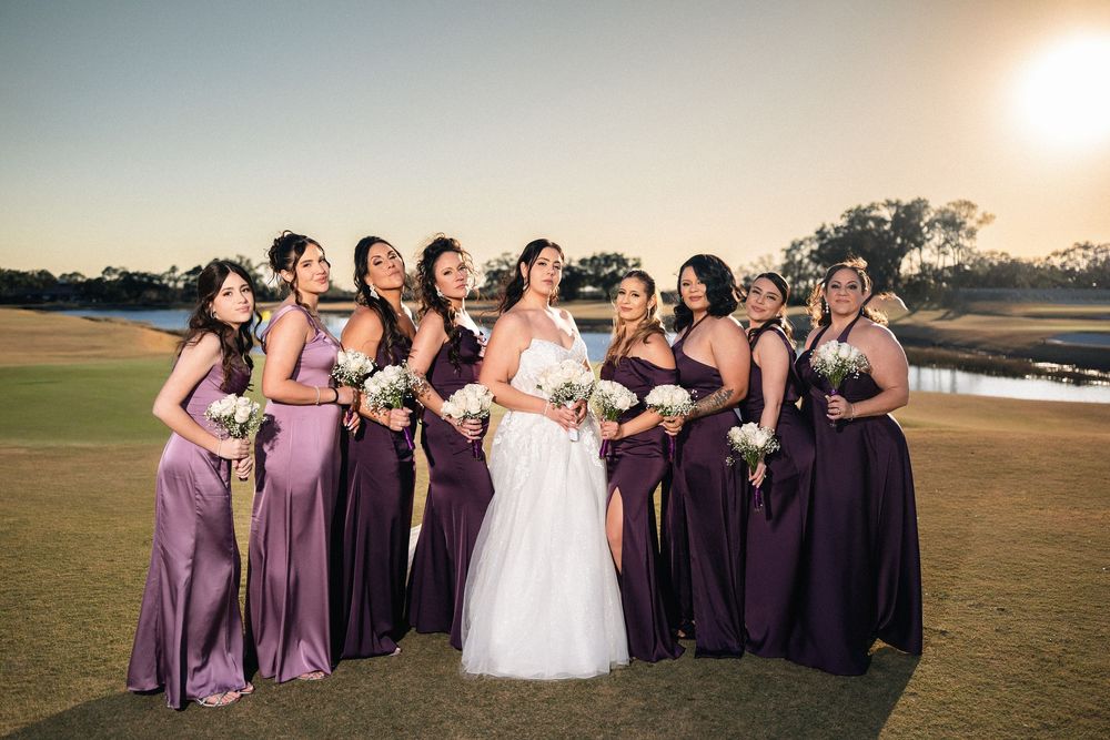 Bride in white gown poses with bridesmaids in purple dresses on golf course at sunset.