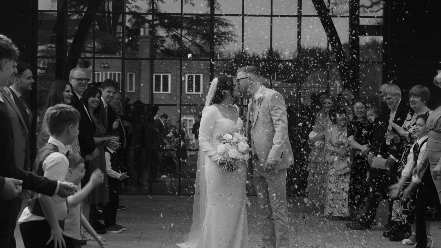 Black and white photo of newlyweds kissing as guests throw confetti outside a wedding venue.