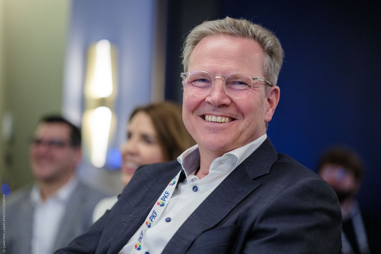 Smiling middle-aged man with glasses in a gray blazer at a professional event.