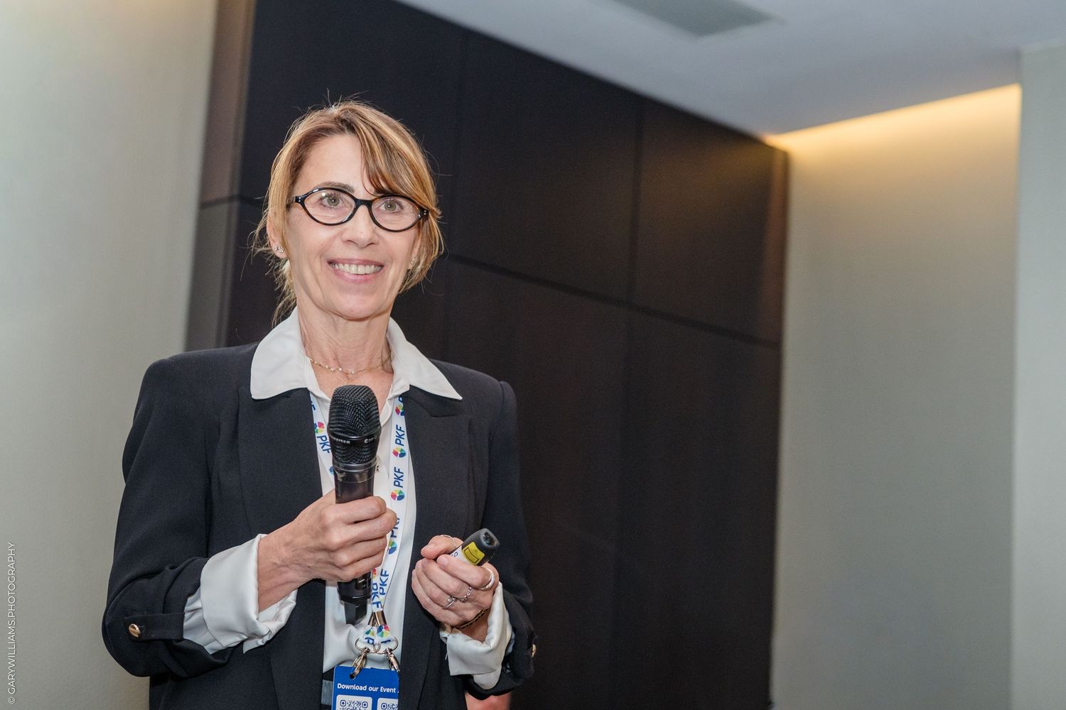 Professional woman in blazer speaking into microphone at corporate conference event.