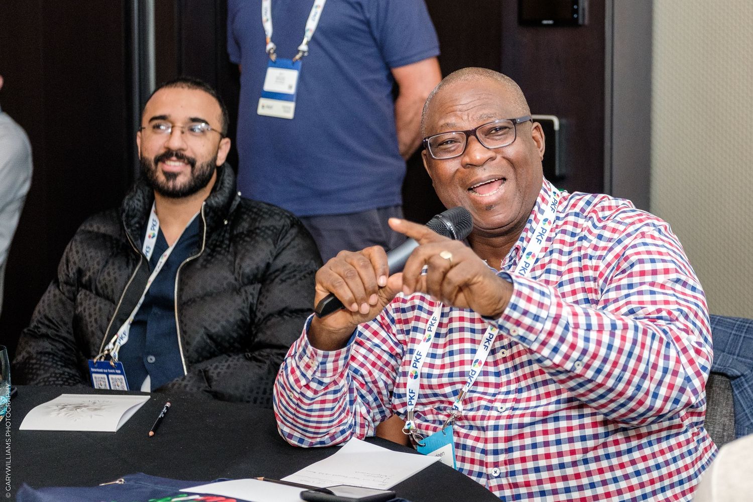 Two men smiling at a conference table, one pointing finger, wearing name badges at a professional event.
