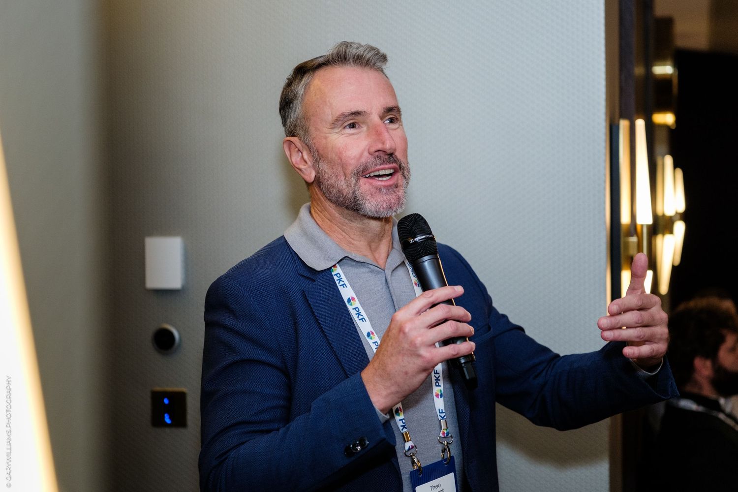 Man in blue blazer speaking into microphone at professional conference event wearing lanyard badge.