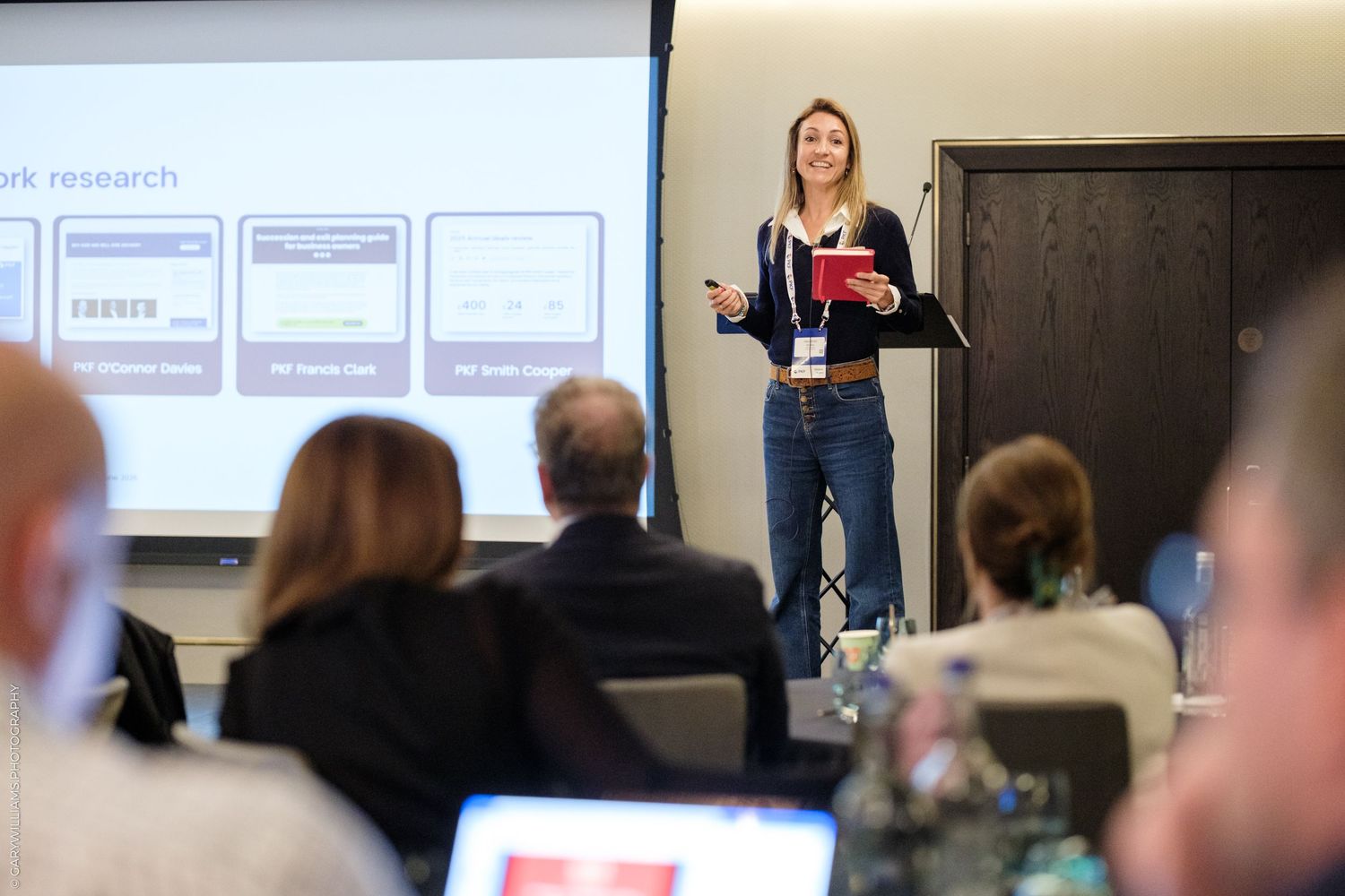 Woman presenting research at a professional conference, standing before a projected slide with an engaged audience.