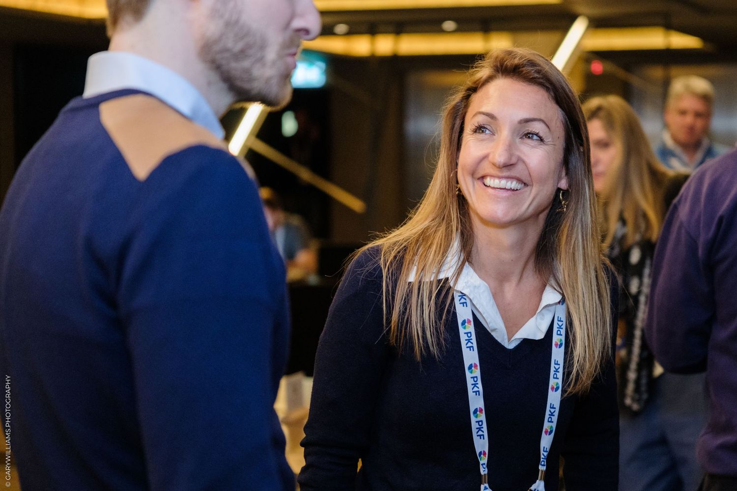 A smiling woman wearing a lanyard engages in conversation at a professional networking event indoors.