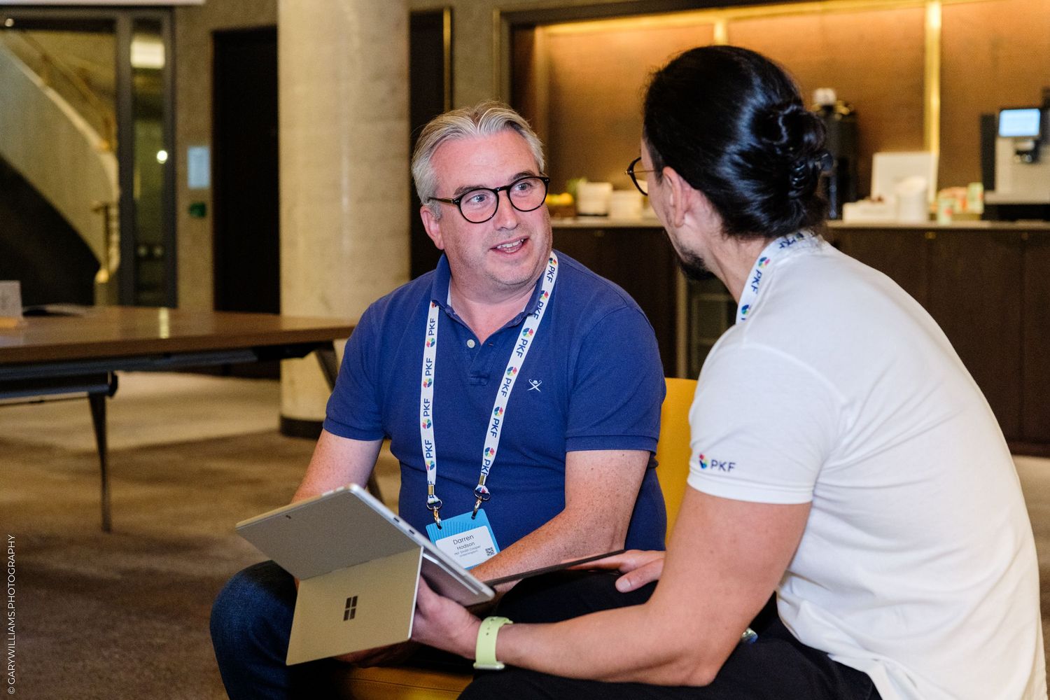 Two professionals engage in conversation at a conference, one holding a tablet, wearing lanyards in a modern venue.