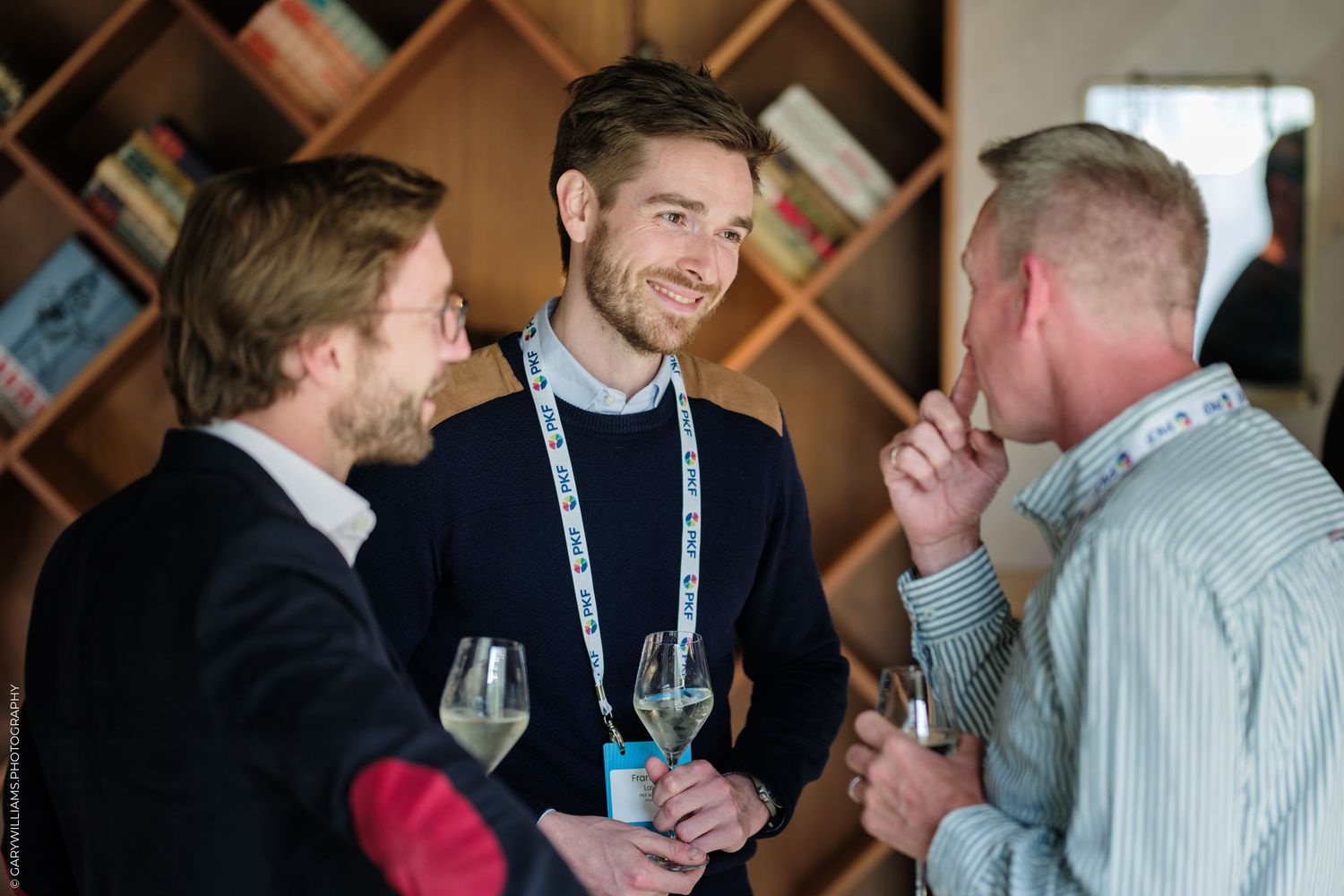Three professionals networking at a conference, holding wine glasses and engaged in conversation near a bookshelf.