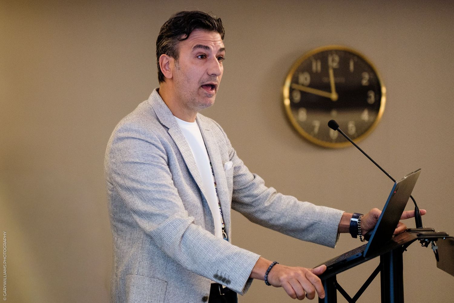 Man in gray blazer speaking at podium with clock on wall behind him at professional conference.