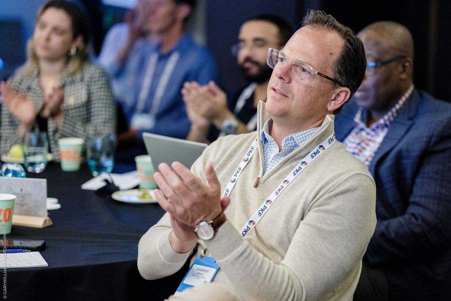 Business professional in beige sweater applauding at corporate conference, holding tablet, wearing lanyard badge.