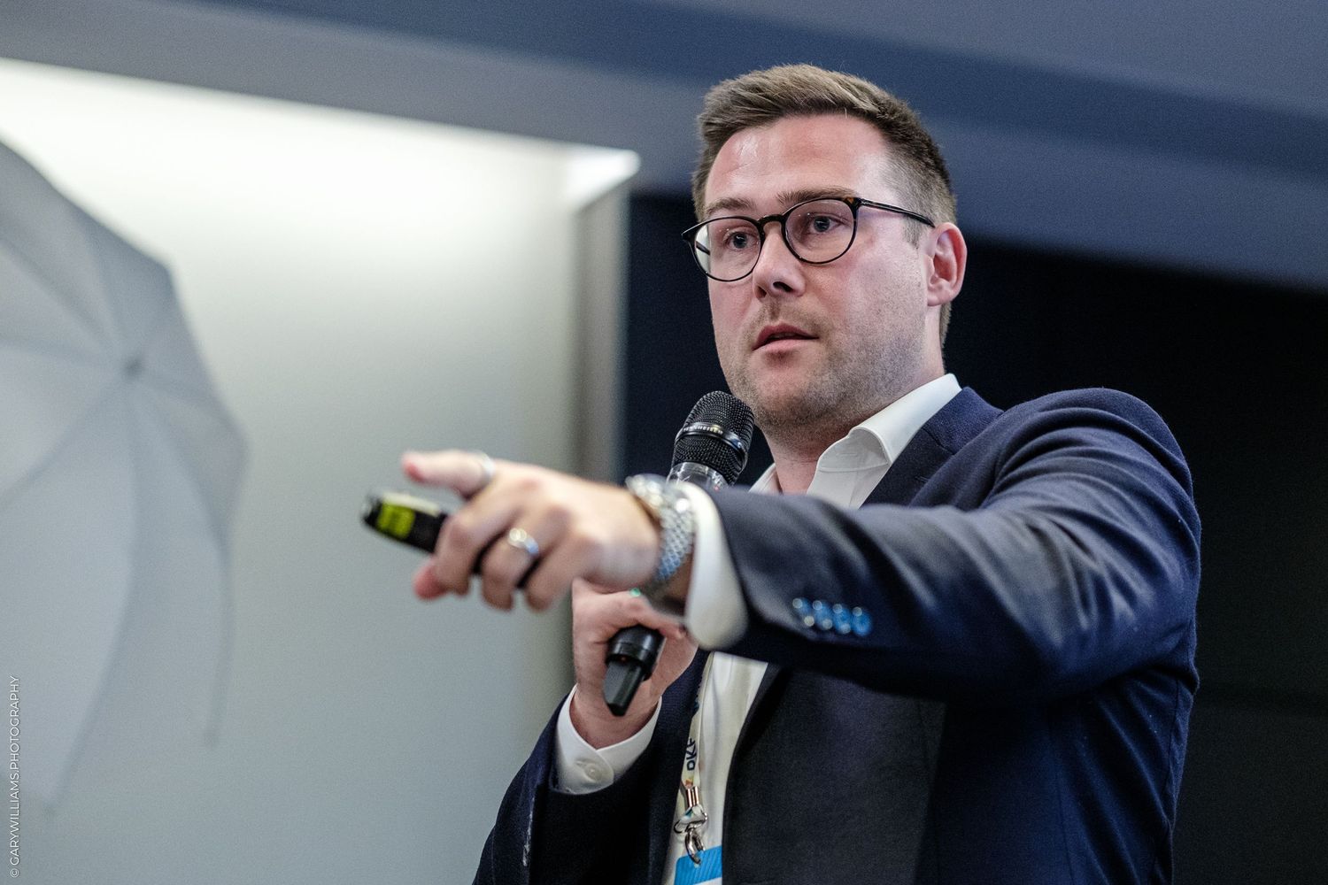 Man in dark suit holding microphone pointing at audience during business presentation.
